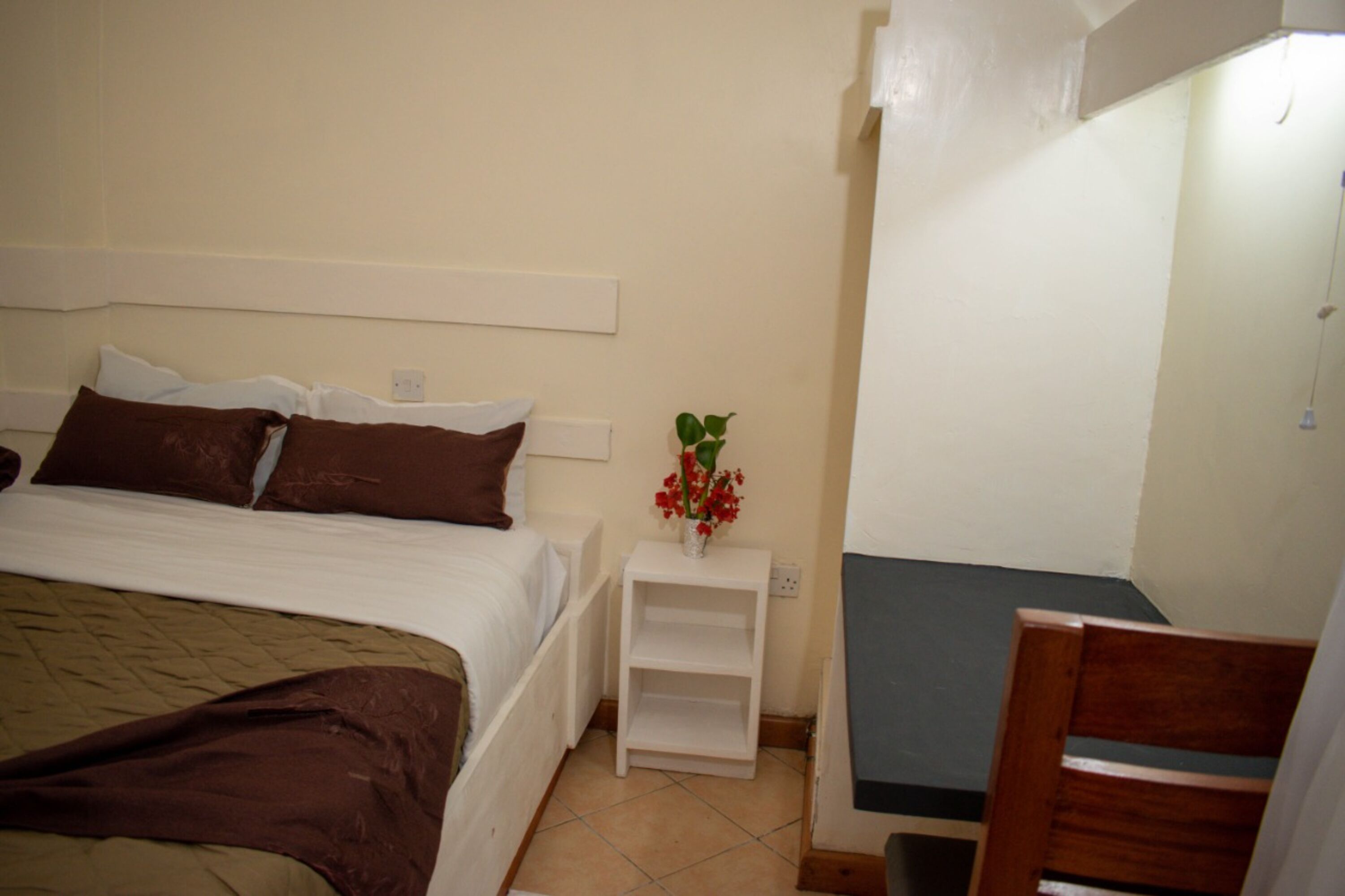 Un dormitorio con una cama, una mesita de noche con un florero, una silla y un escritorio pequeño.