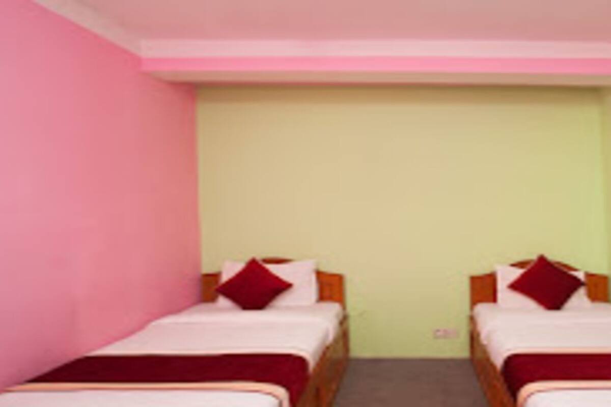 Quarto com duas camas de solteiro, cada uma com almofada vermelha e colcha branca. As paredes são pintadas em tons de verde claro e rosa, e há uma escada de madeira encostada na parede.