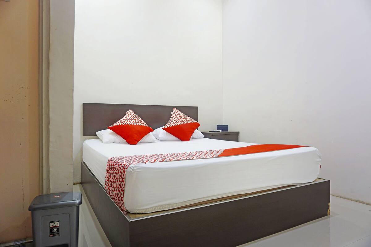 Una cama bien hecha con ropa blanca y roja, un cabecero gris y una mesita de noche con una lámpara.