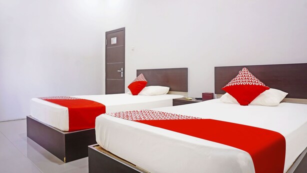 A hotel room with two beds, each with a red and white bedspread and matching pillows. There is a wardrobe and a nightstand with a lamp.