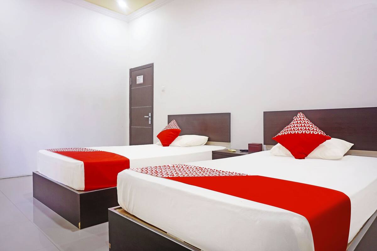 Habitación de hotel con dos camas, cada una con cubrecama rojo y blanco y almohadas a juego. Se aprecia un armario y una mesita de noche con lámpara.