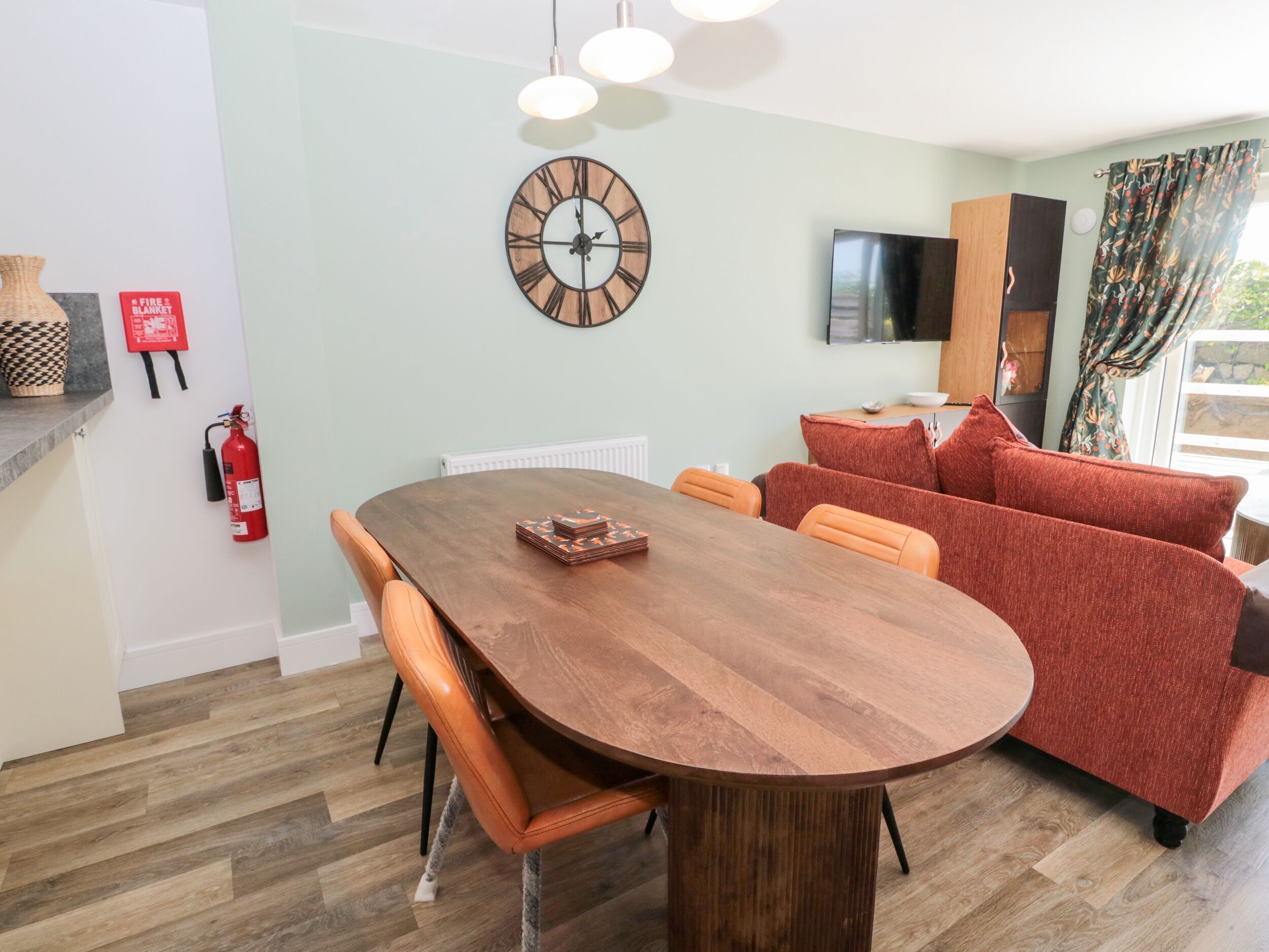 A dining area with a wooden table, orange chairs, a red sofa, a wall clock, a flat-screen TV, and a fire extinguisher.