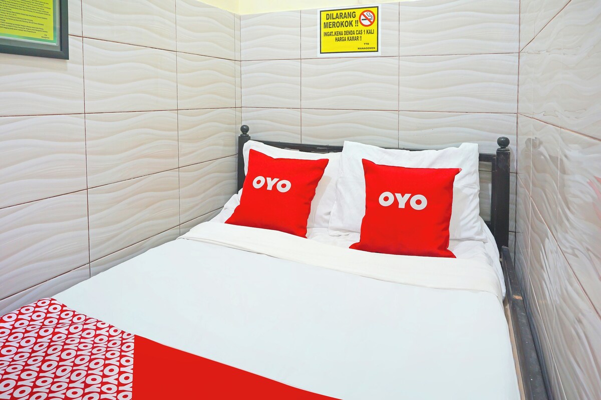 A hotel room with a bed, two red pillows with the word 'OYO' on them, a framed sign on the wall, and a patterned blanket.