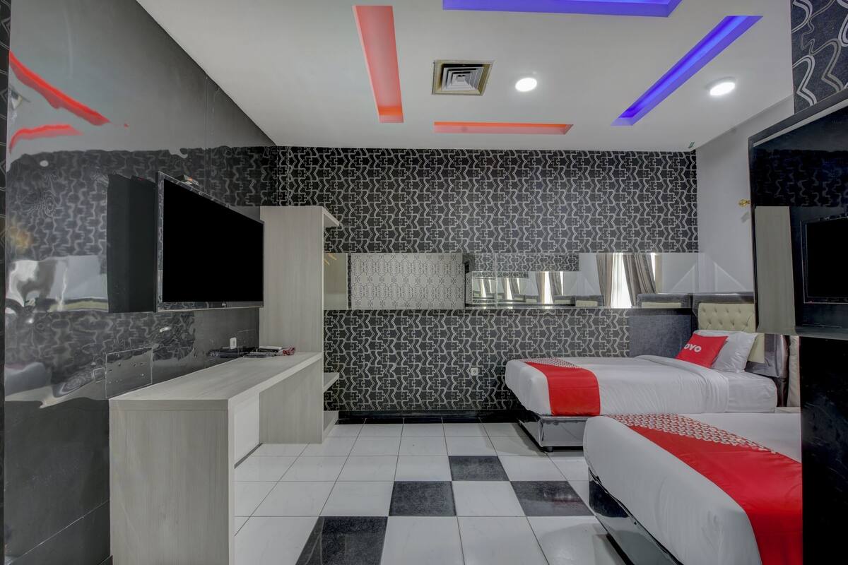 A modern hotel room with a checkered floor, a kitchenette, a TV, and two beds with red accents.