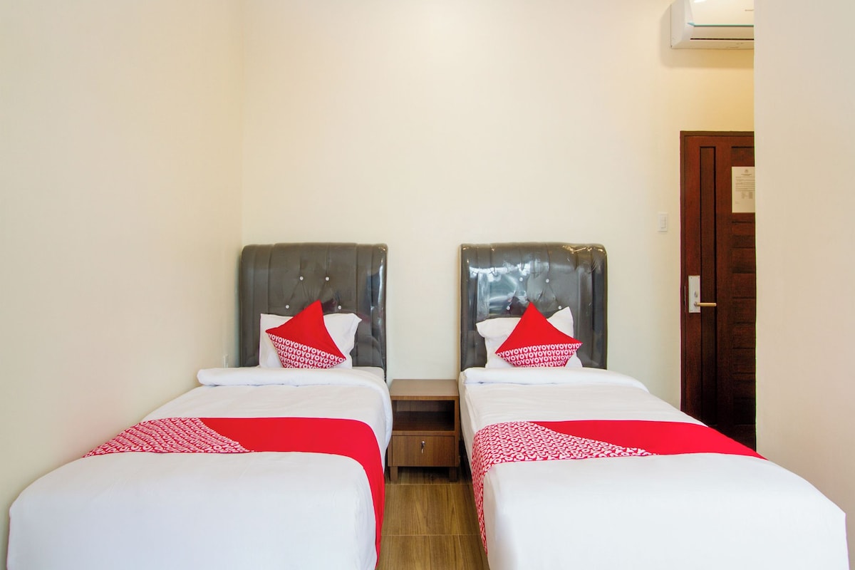 Une chambre d’hôtel avec deux lits simples, chacun agrémenté d’un oreiller rouge et blanc, d’une table de chevet en bois et d’une porte avec une pancarte.
