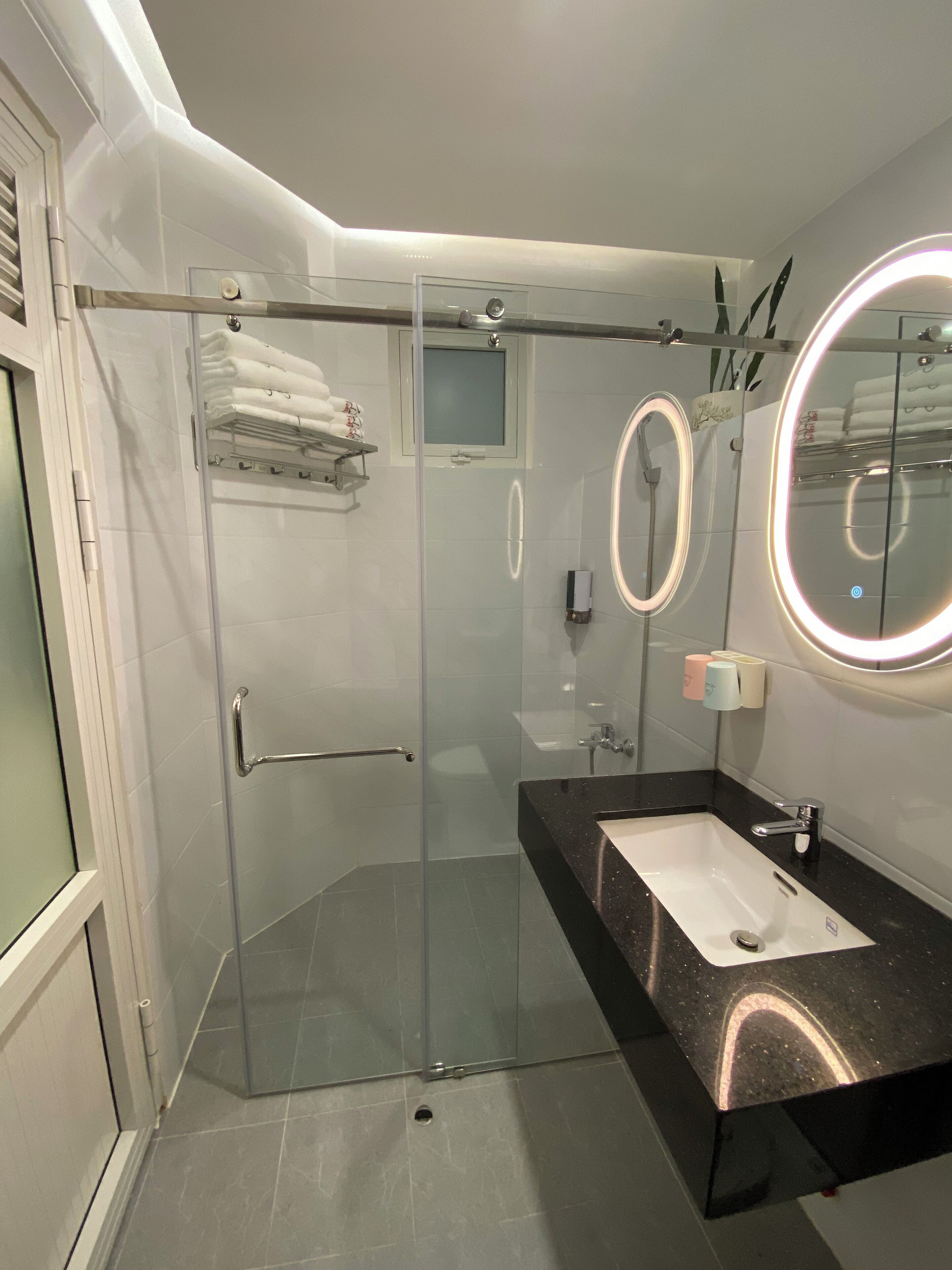 A modern bathroom with a glass-enclosed shower, a vanity with a black countertop, and a round mirror with a lighted frame.