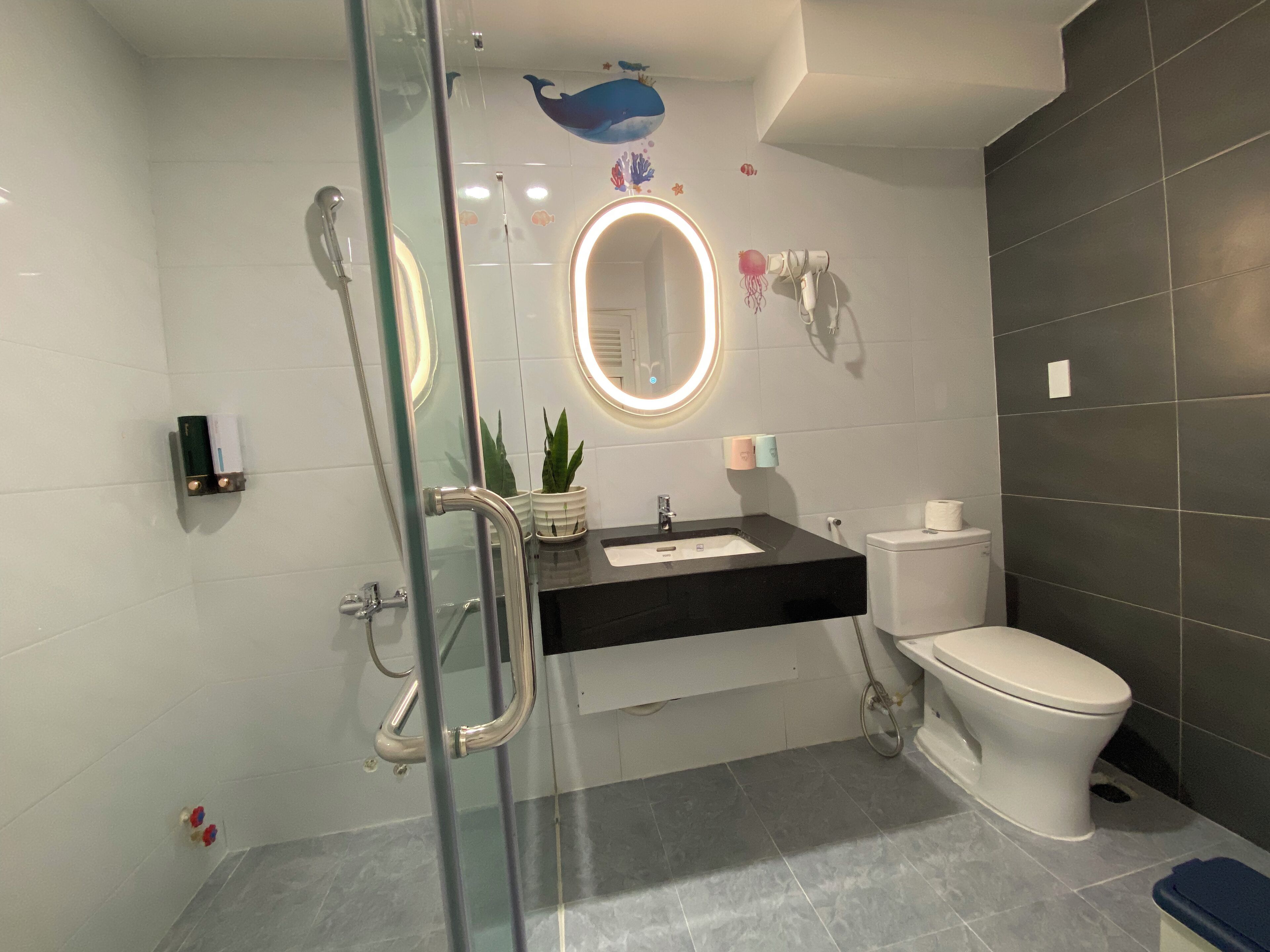 A modern bathroom with a glass shower enclosure, a black sink, a white toilet, and a round mirror with a light.