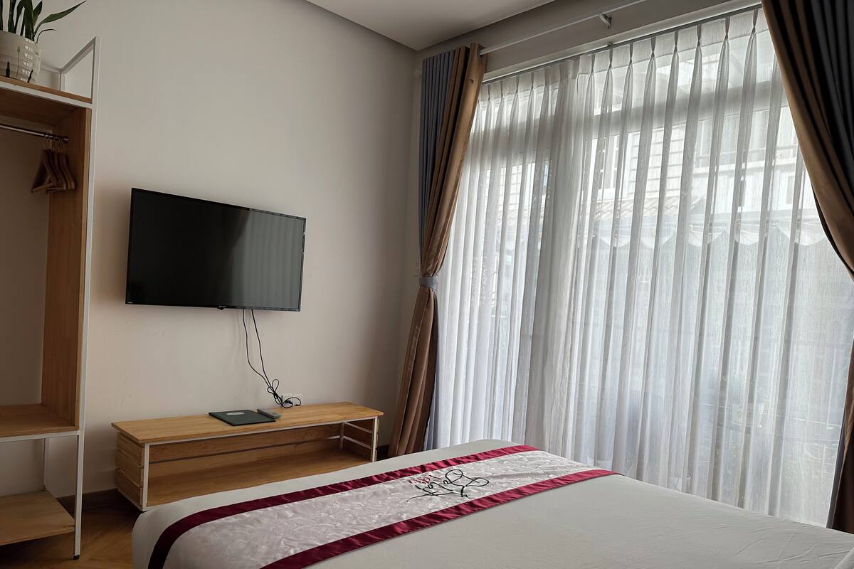 A hotel room with a bed, a TV mounted on the wall, a wooden desk, and a window with sheer curtains.