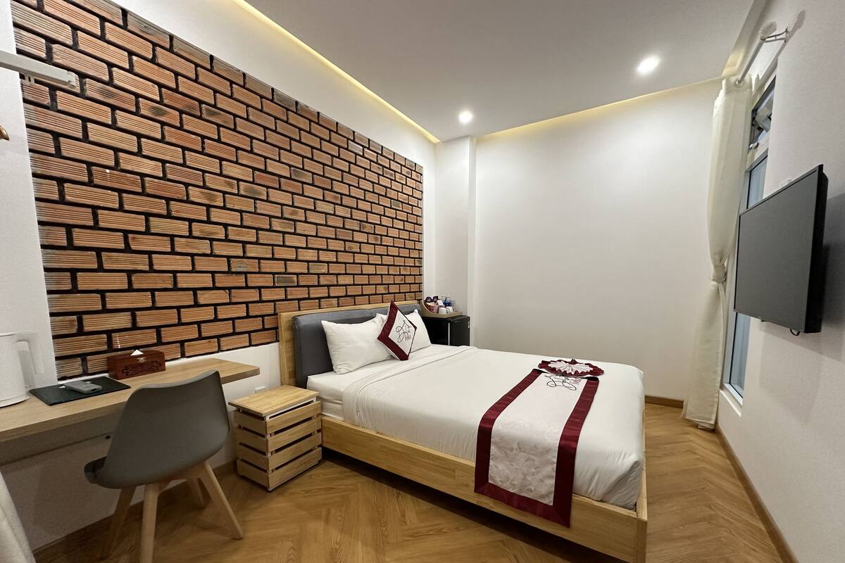 A modern bedroom with a brick-patterned wall, a wooden bed with a red and white bedspread, a desk with a chair, and a TV mounted on the wall.