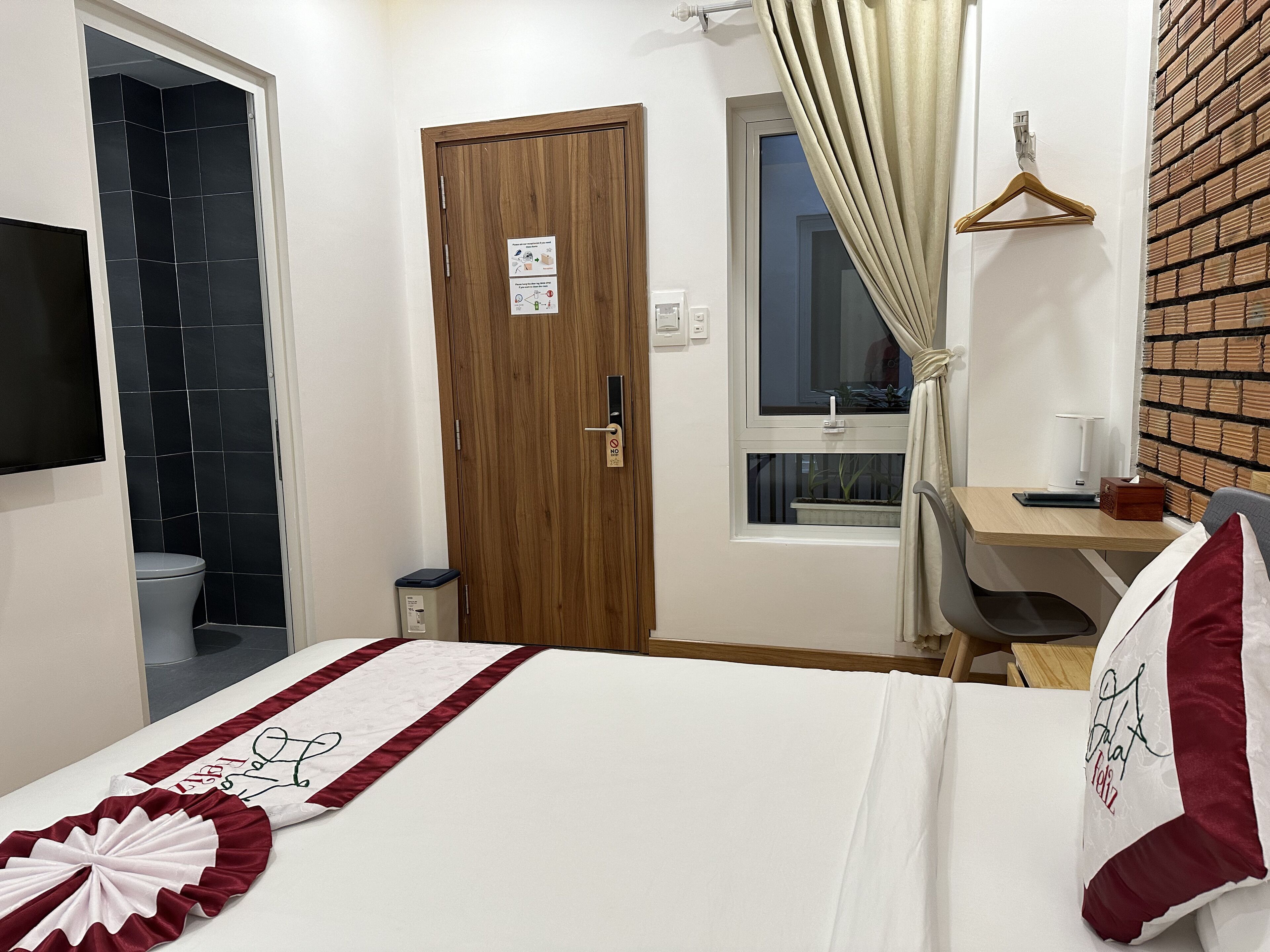 A hotel room with a bed, a wooden door, a window with curtains, a desk with a chair, and a TV mounted on the wall.