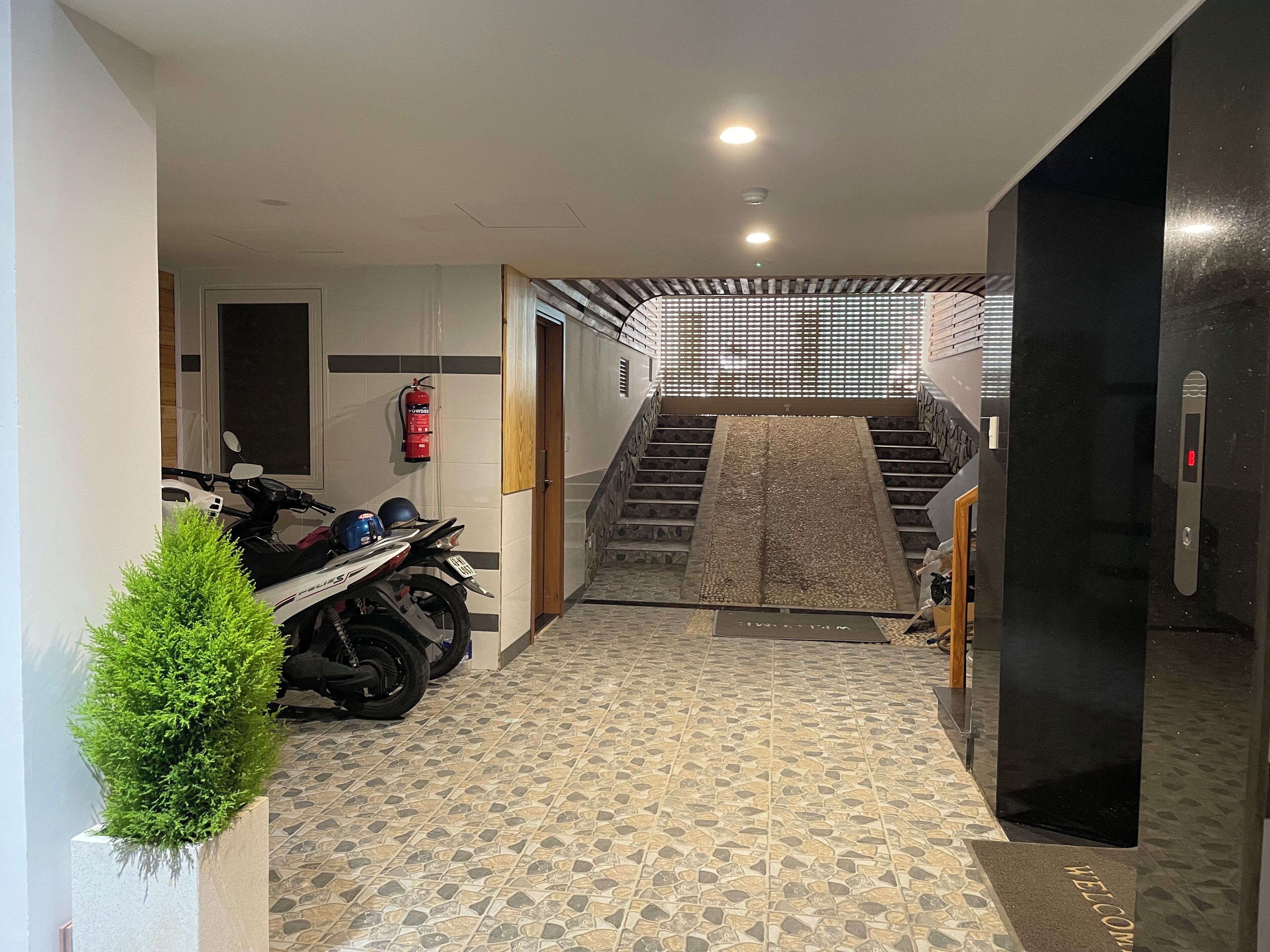 Un couloir avec un sol carrelé, un extincteur, une moto et un escalier menant à un étage supérieur.