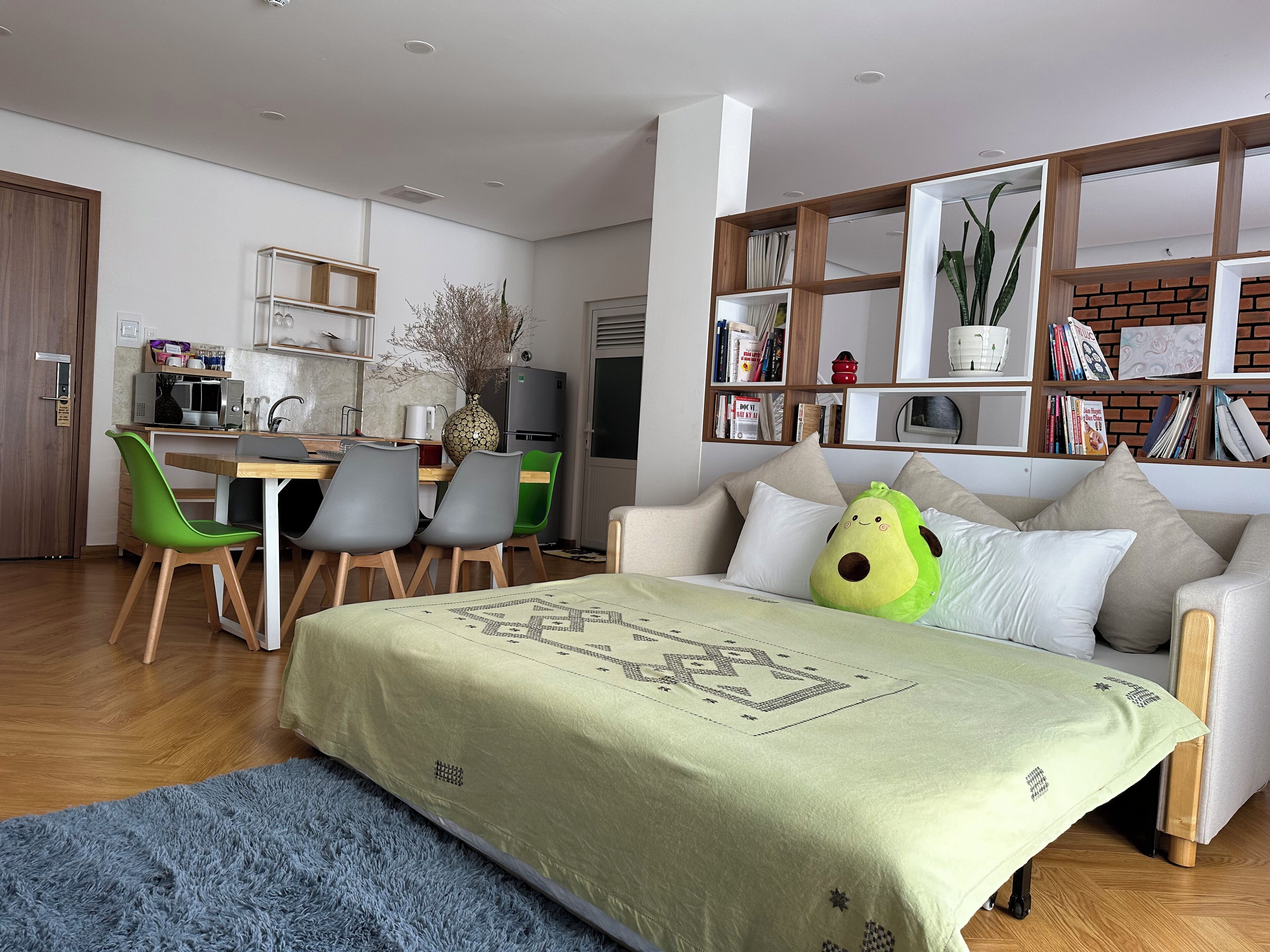 A modern living room with a sofa, a dining area with chairs, a kitchen with appliances, and a bookshelf.