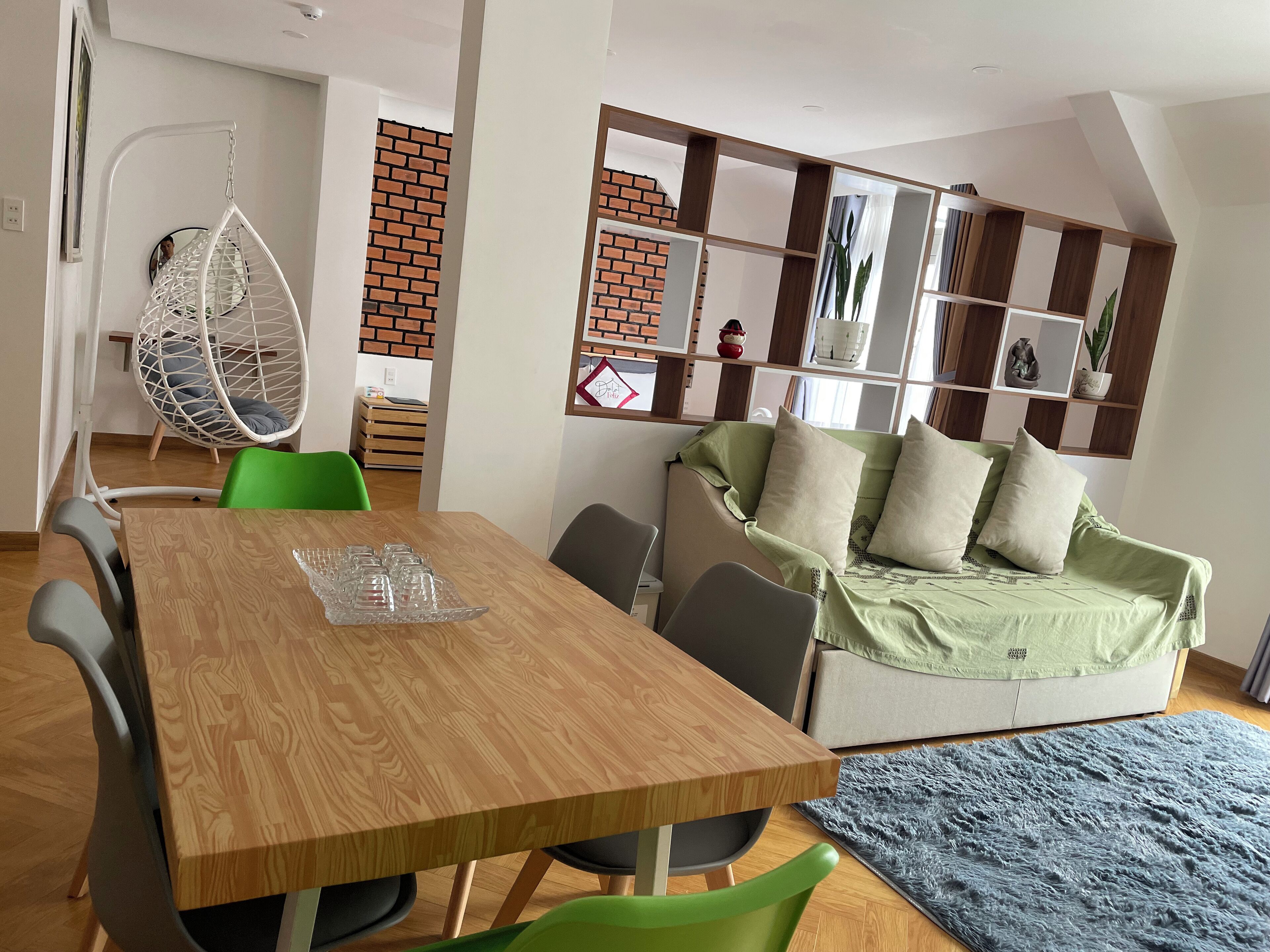 Una sala de estar moderna con una mesa de comedor de madera, sillas verdes, un sillón colgante, un sofá con cojines y una pared de ladrillo.