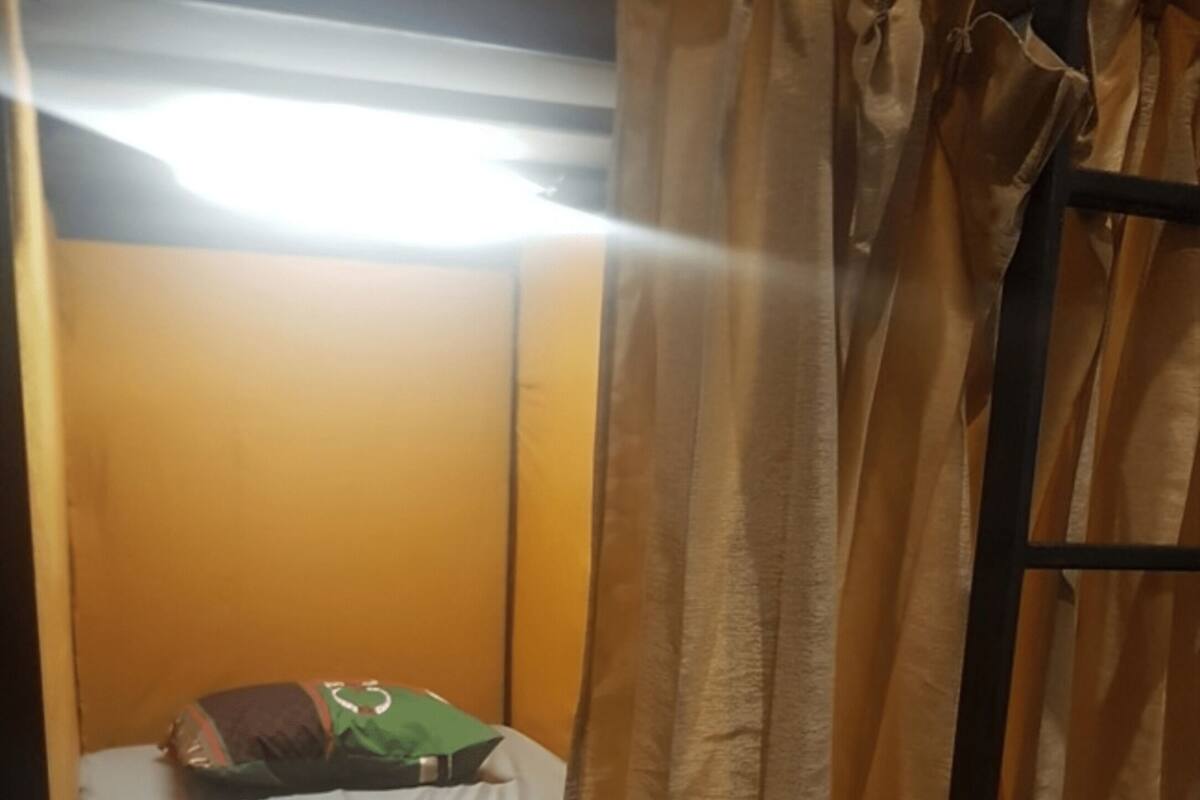 Un lit simple avec un drap blanc et un oreiller vert, une tête de lit en bois et un rideau tiré partiellement sur le côté.
