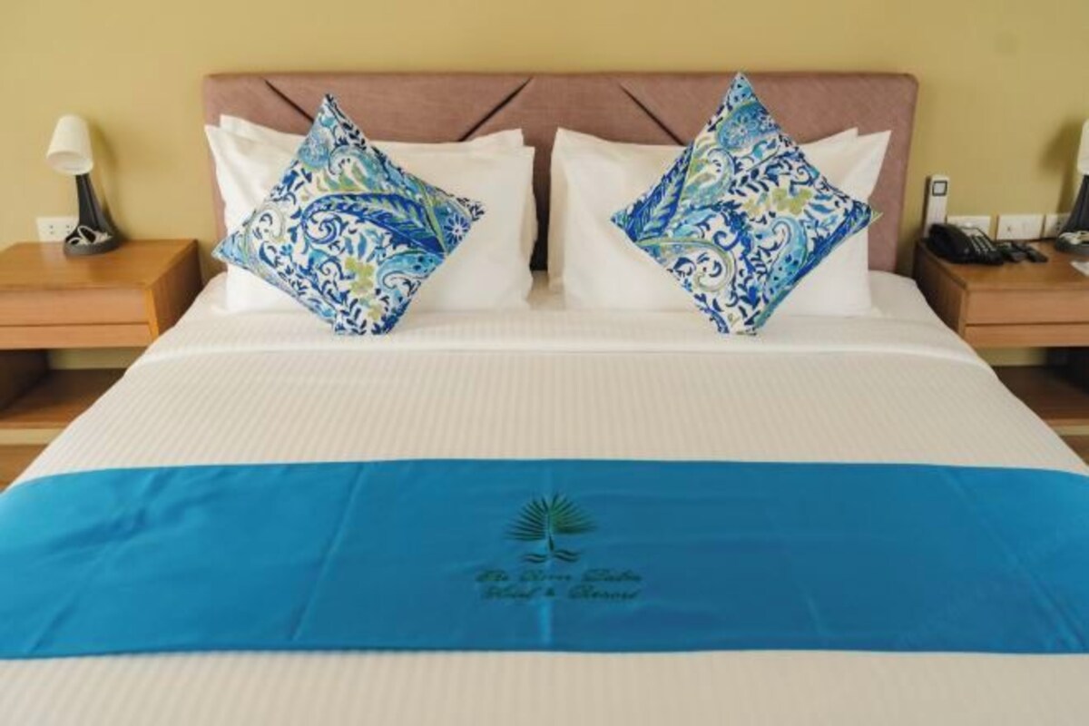 Una cama con colcha azul, dos almohadas decorativas, una mesita de noche con lámpara y un teléfono a la derecha.