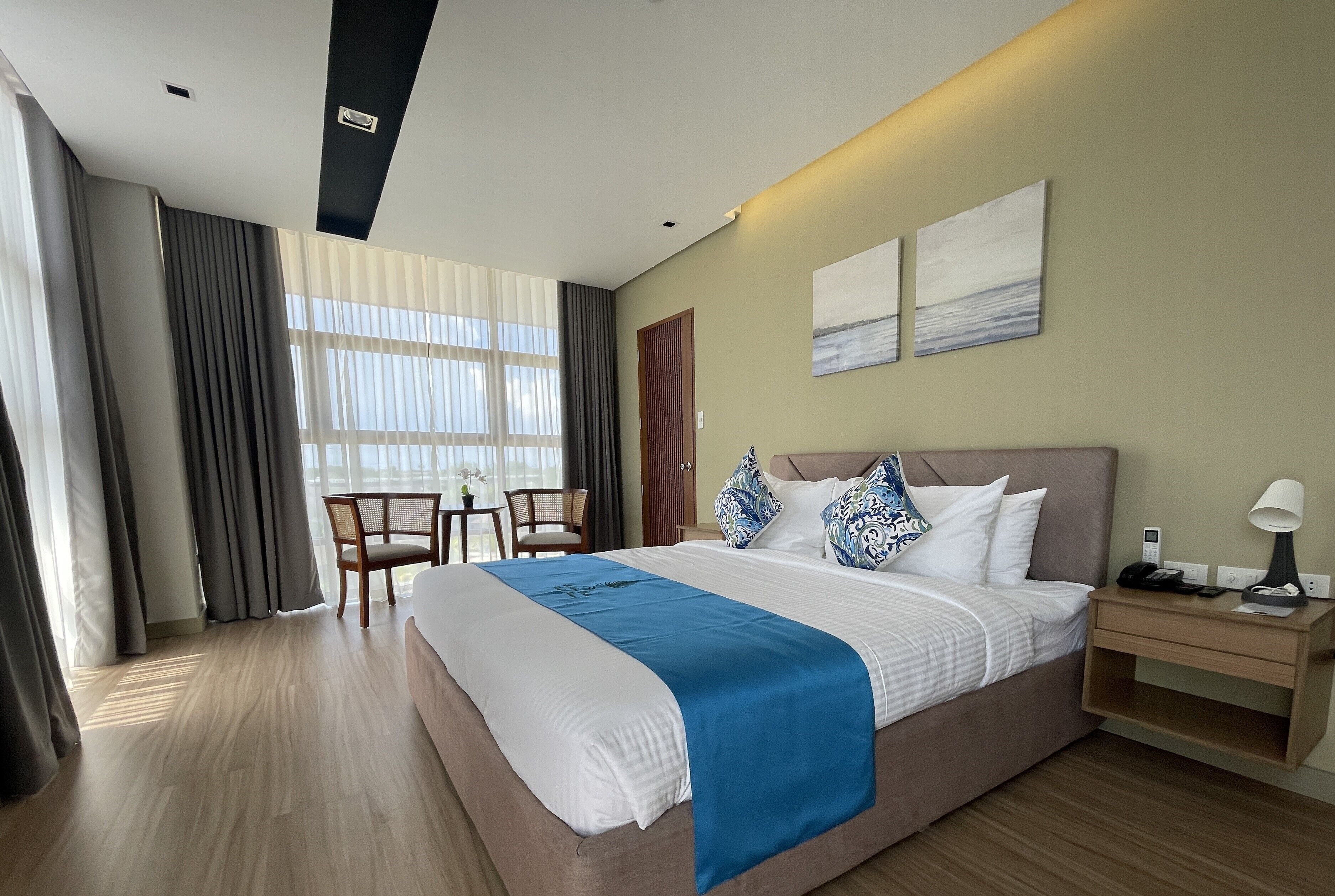 A hotel room with a large bed, a desk, and a chair. There are curtains, a window, and artwork on the wall.