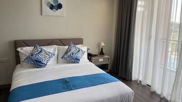 Una cama bien hecha con colcha azul y blanca, dos almohadones decorativos, una mesita de noche con lámpara y una ventana con cortinas de encaje.