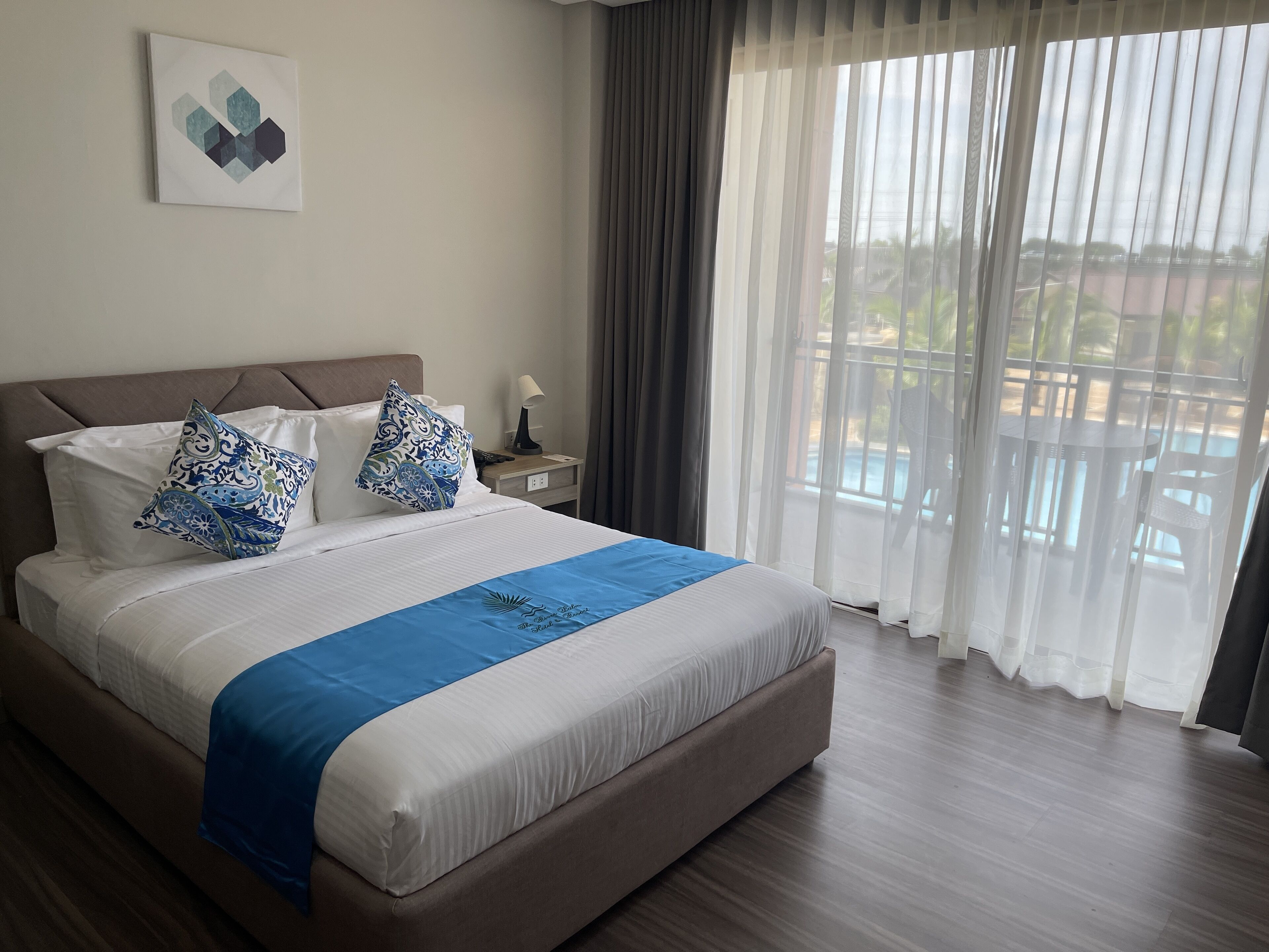 Ein Hotelzimmer mit einem großen Bett, einem Nachttisch mit Lampe und Blick auf einen Pool durch transparente Vorhänge.