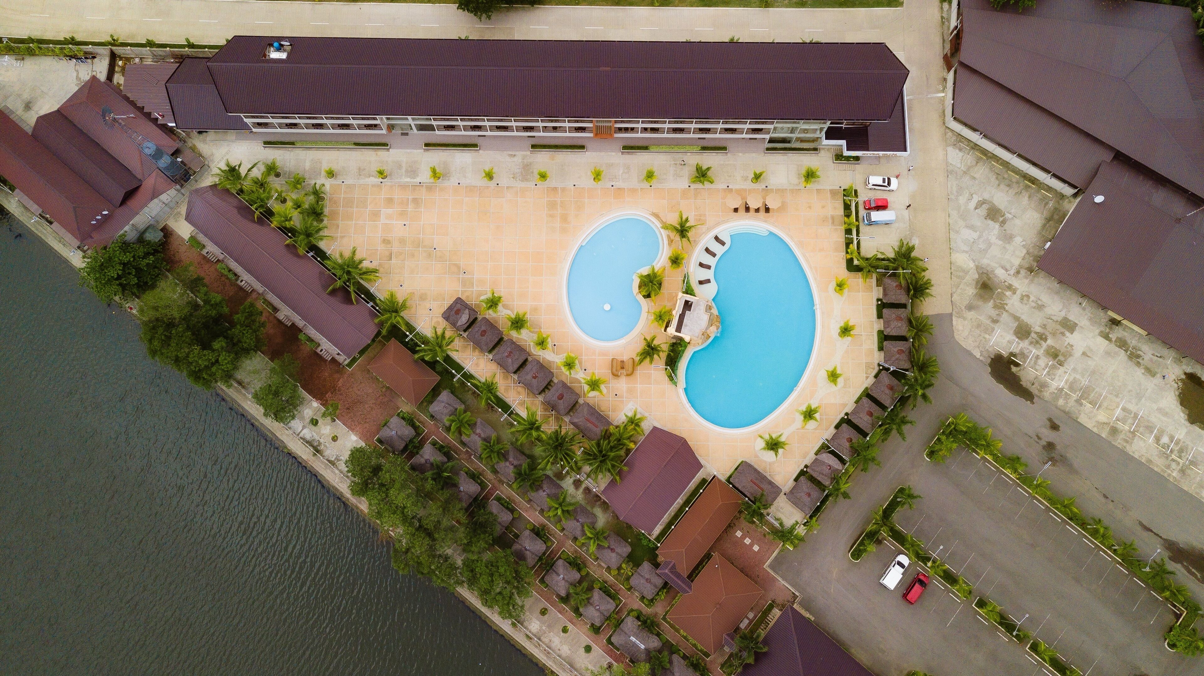 Vista aérea de un resort con dos piscinas, un paseo cubierto y un edificio grande con techo oscuro.