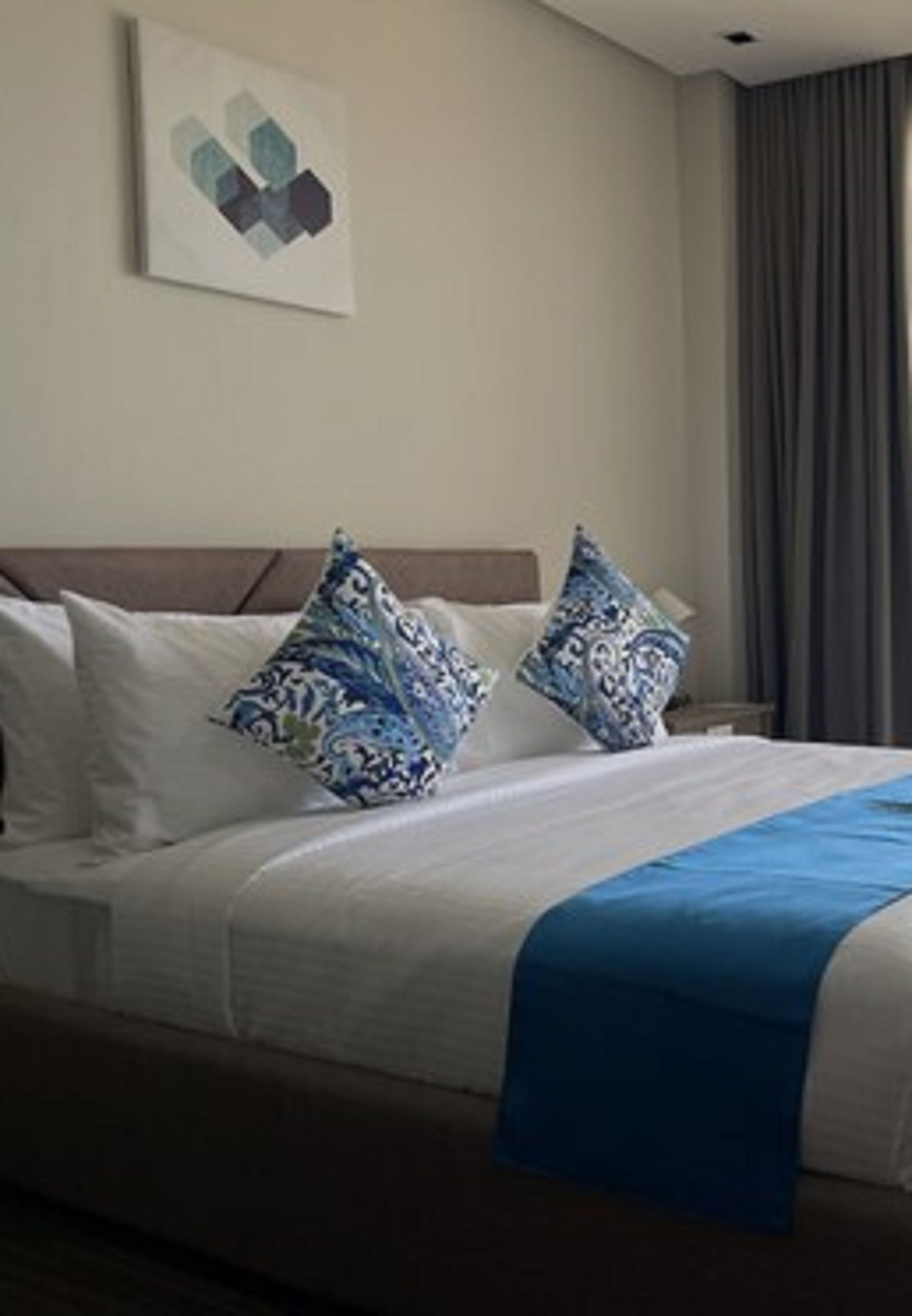 A hotel room with a bed, pillows, a blue blanket, and a painting on the wall.