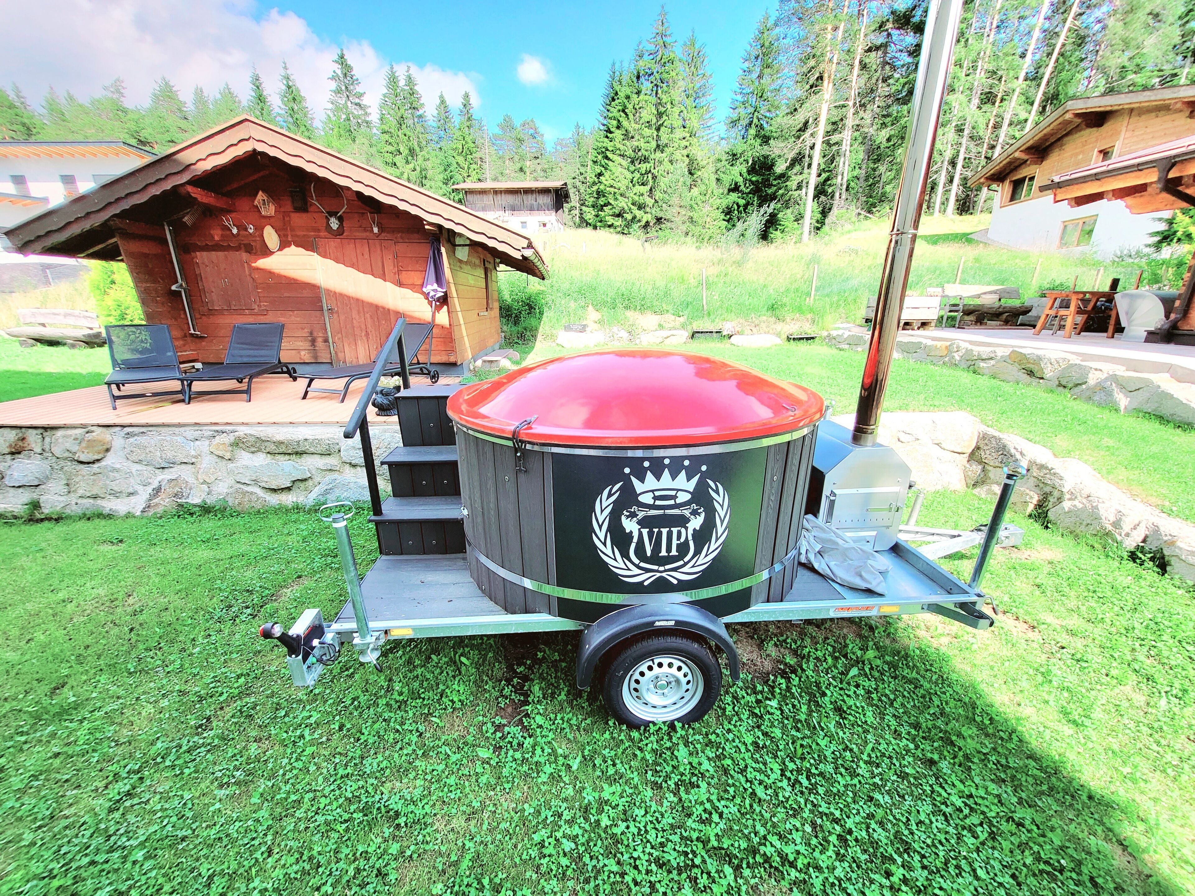 A portable VIP hot tub with a red cover, a metal frame, and a logo, set on a grassy area with a wooden deck and chairs in the background.