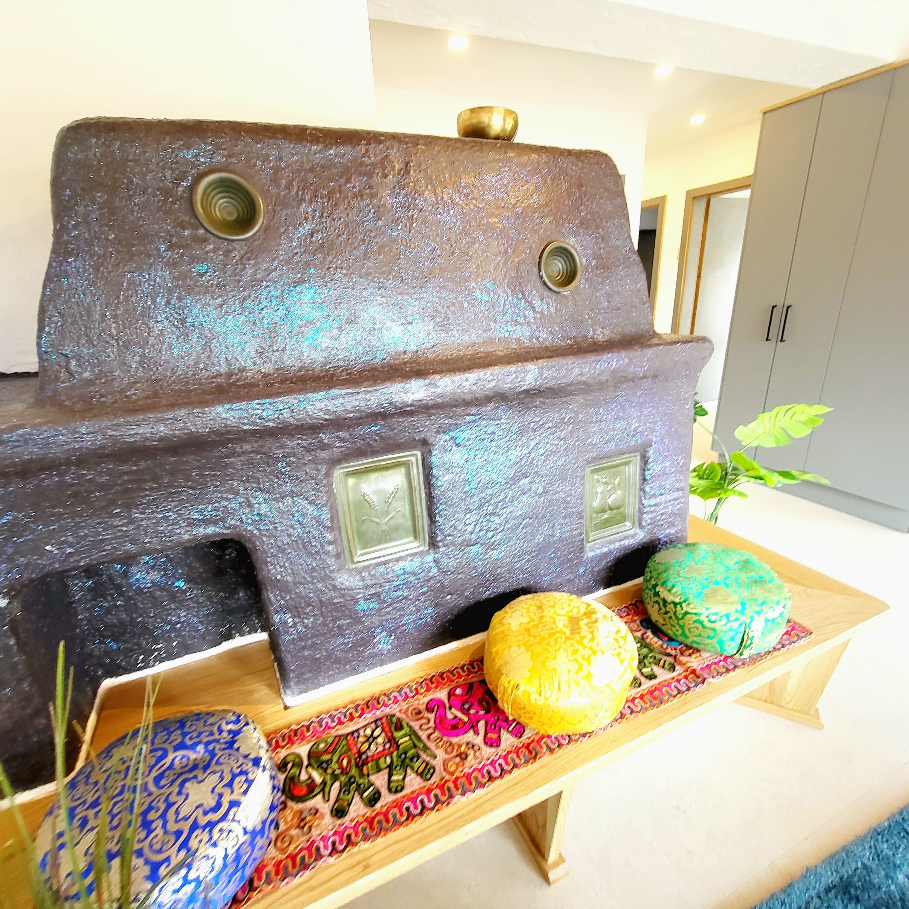 Een decoratief metalen beeldhouwwerk, dat een huis met twee ramen en een deur lijkt, geplaatst op een houten tafel met kleurrijke geweven matten en decoratieve objecten.