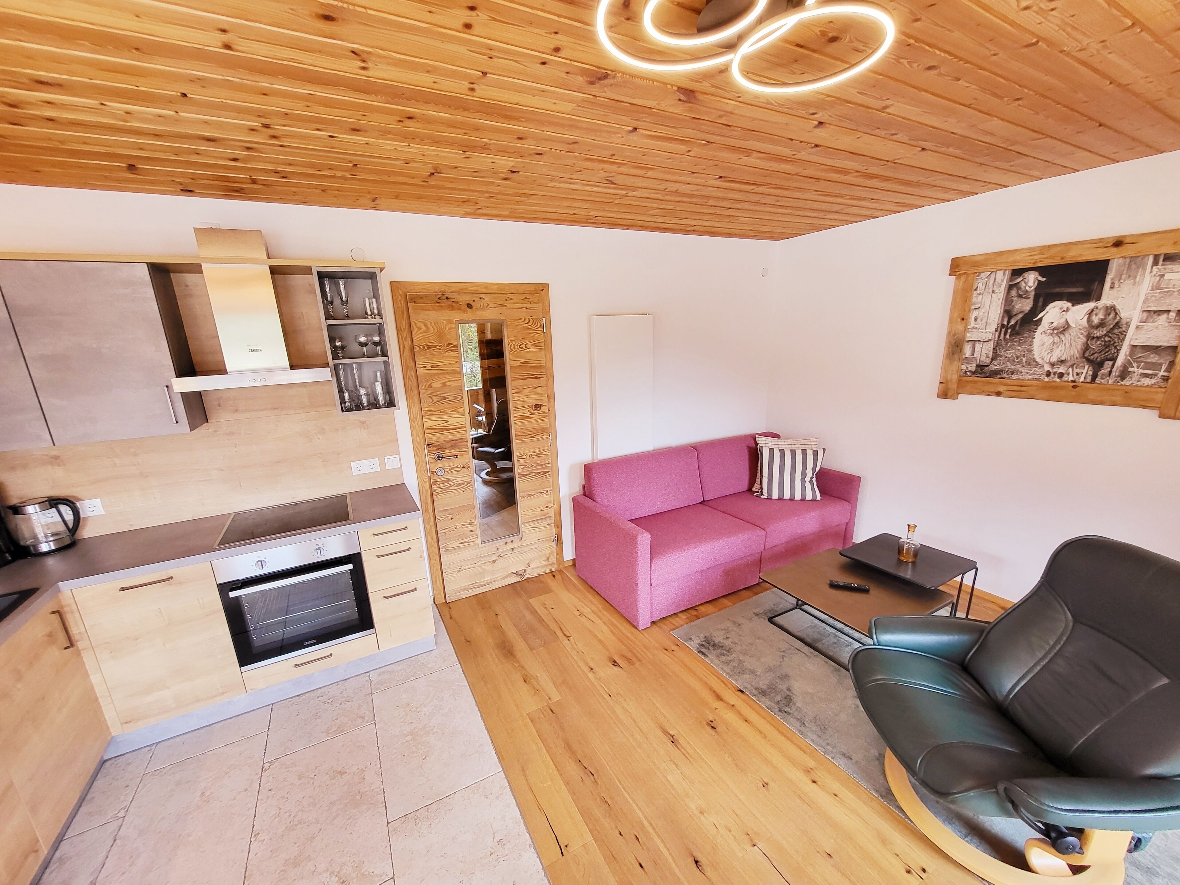Una cocina moderna con armarios de madera y horno empotrado, un sofá rosa, un sillón de cuero negro y un cuadro en la pared.