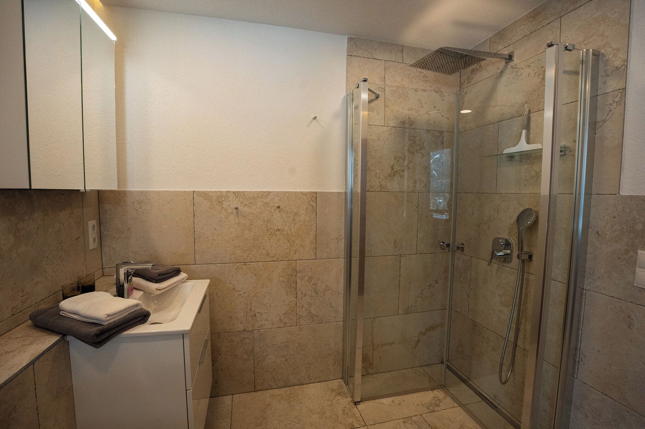 Ein modernes Badezimmer mit einer gläsernen Duschkabine, einem weißen Waschbecken und ordentlich zusammengelegten Handtüchern.