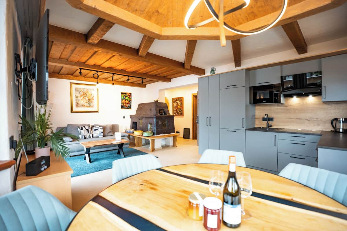 Una cocina moderna con armarios grises, un horno integrado y un microondas. Hay una zona de comedor con una mesa y sillas de madera, y un sofá al fondo.