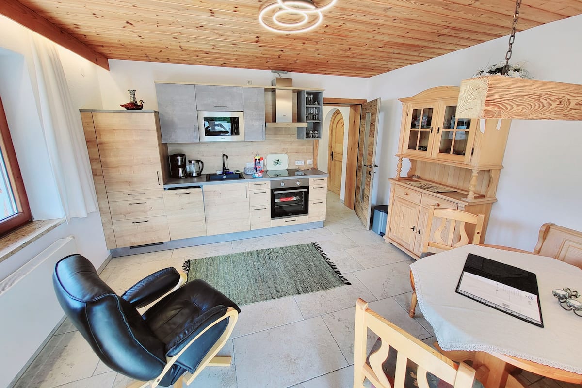 Una cocina moderna con armarios de madera, microondas integrado y cafetera. Hay un comedor con una mesa y sillas, y una computadora portátil sobre la mesa.