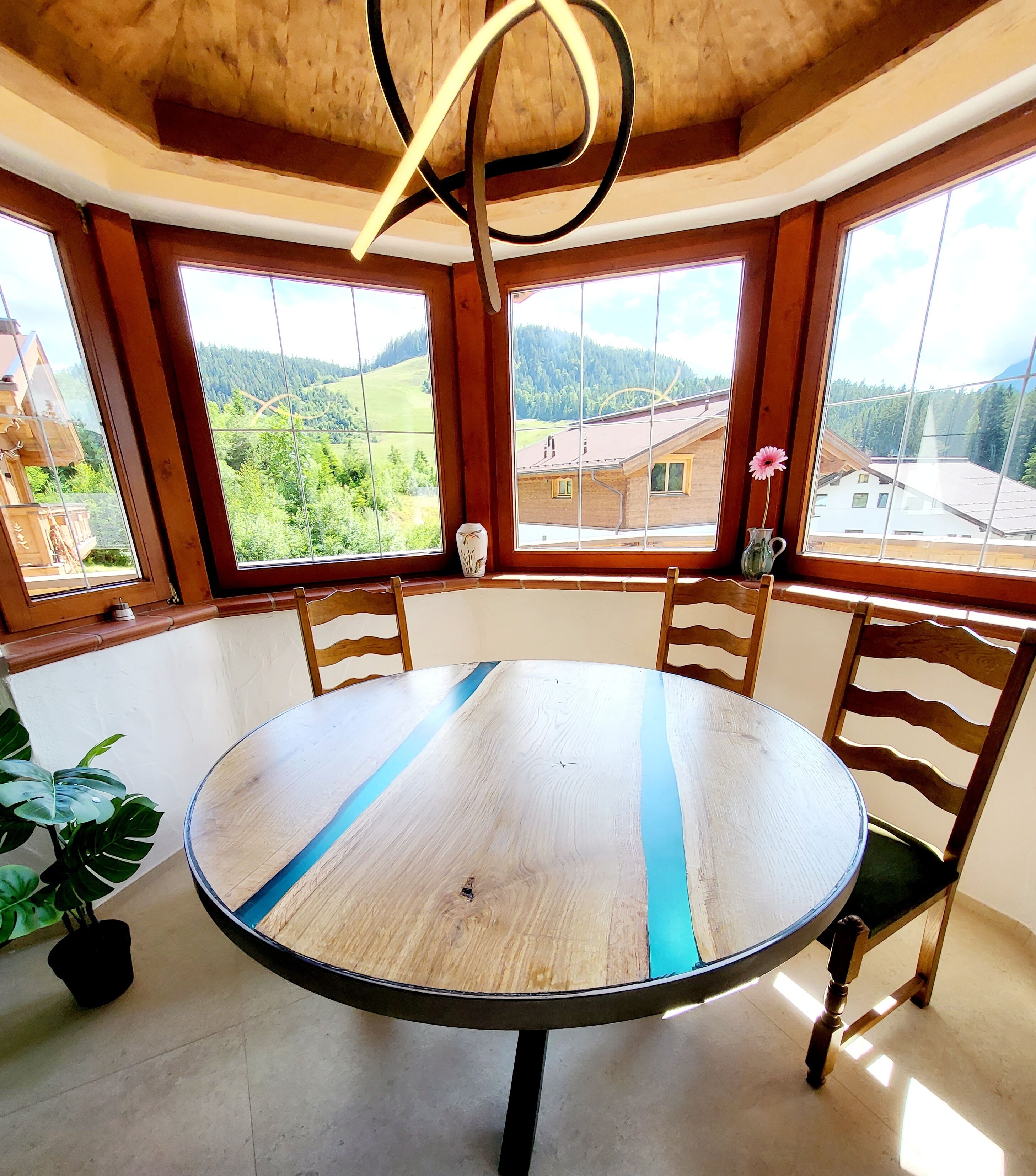 Una mesa redonda de madera con sillas, ventanas amplias con vistas a un paisaje verde, y una lámpara colgante.