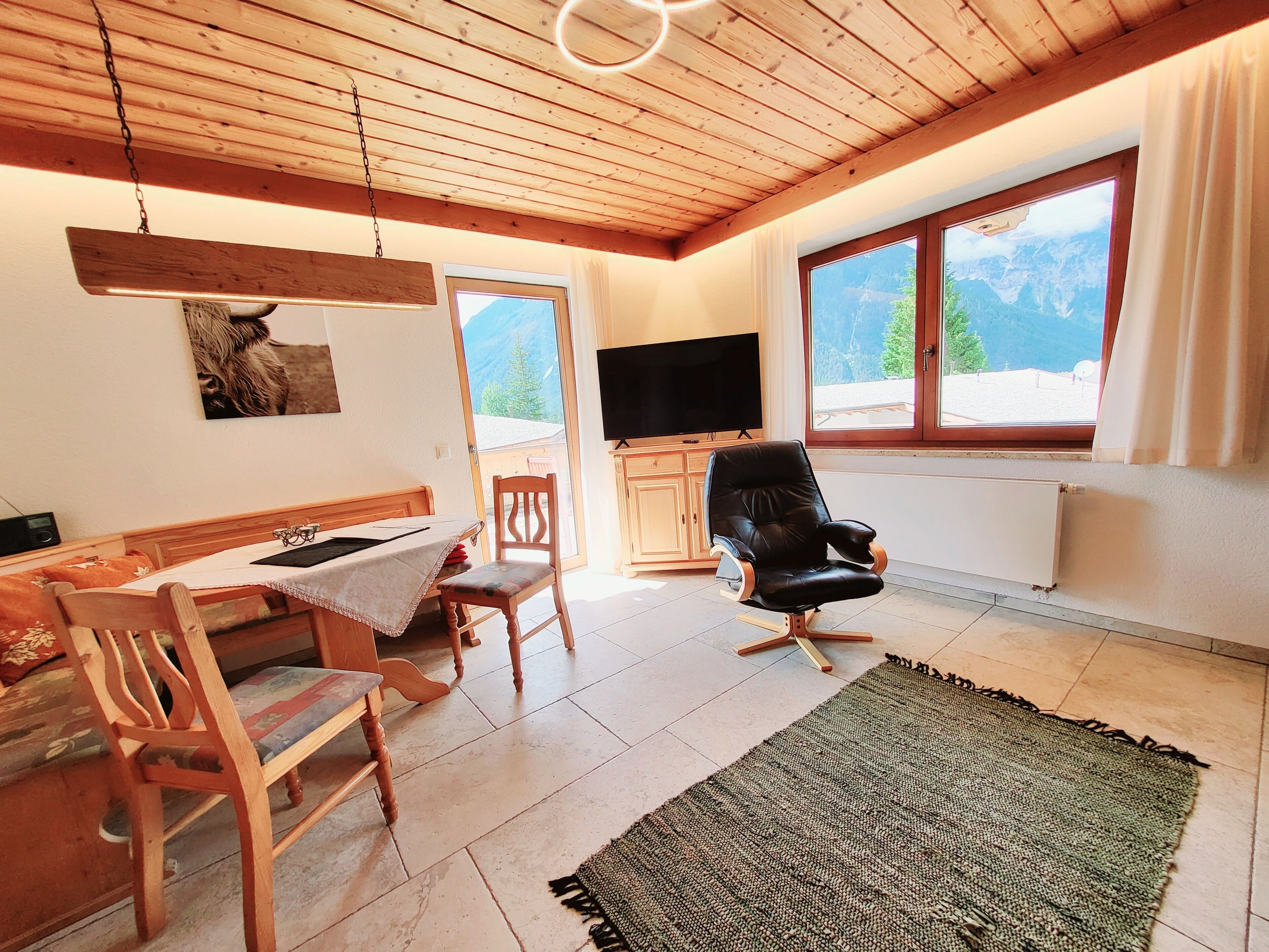 A cozy living room with a wooden ceiling, a television, a black leather chair, a wooden dining table with chairs, and a view of mountains through the windows.