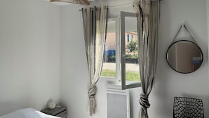 A bedroom with a bed, a bedside table, a hanging light fixture, a window with curtains, and a wall-mounted mirror.