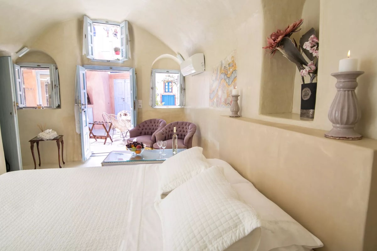 A bedroom with a bed, a table with a fruit bowl, and a chair. There are two windows with open shutters, and a wall-mounted air conditioner.