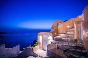 A coastal building with a stone staircase leading to a door, overlooking a body of water. - Kastro Oia Houses  (Santorini)