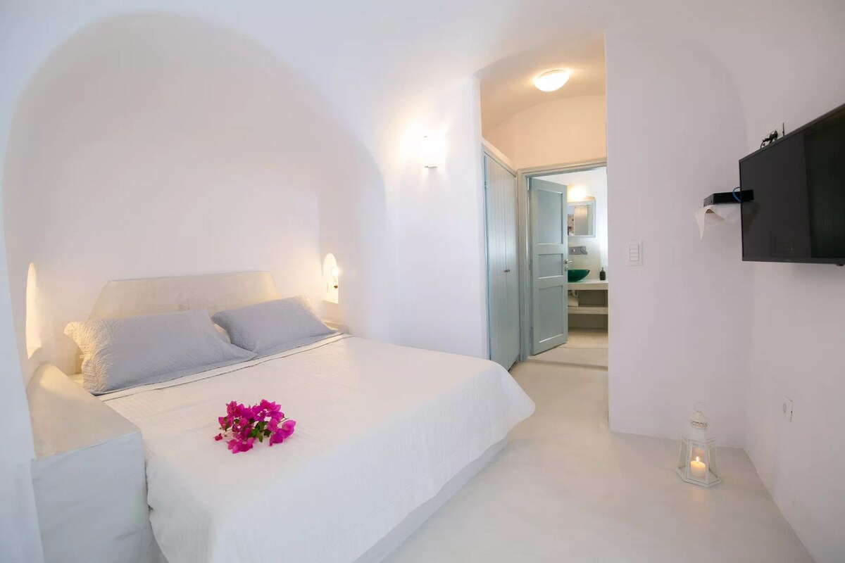 A bedroom with a bed, a nightstand, a wall-mounted TV, and a bathroom visible through an open door.