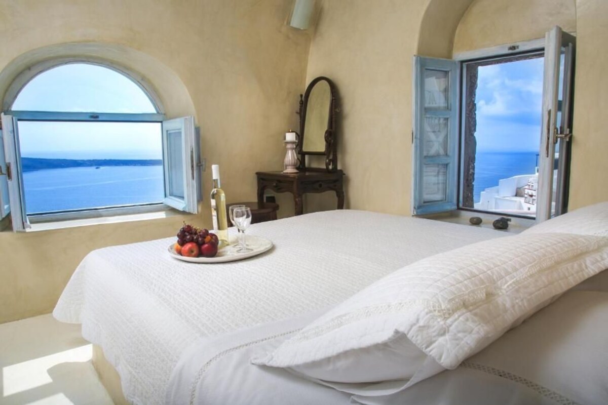 A bedroom with a bed, a bedside table with a plate of fruit, a bottle, and a glass, and a view of the sea through an open window.
