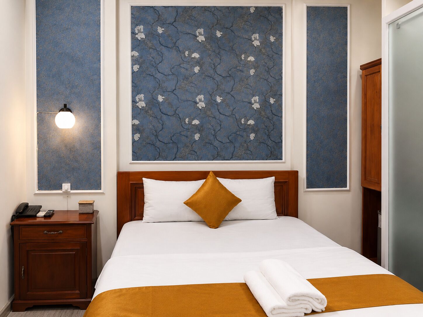 Une chambre d’hôtel avec un lit, une table de chevet avec un téléphone, une lampe fixée au mur et une grande œuvre d’art florale au-dessus du lit.