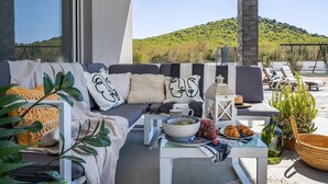 Un área de estar moderna al aire libre con un sofá, cojines y una mesa dispuesta con frutas y una botella.