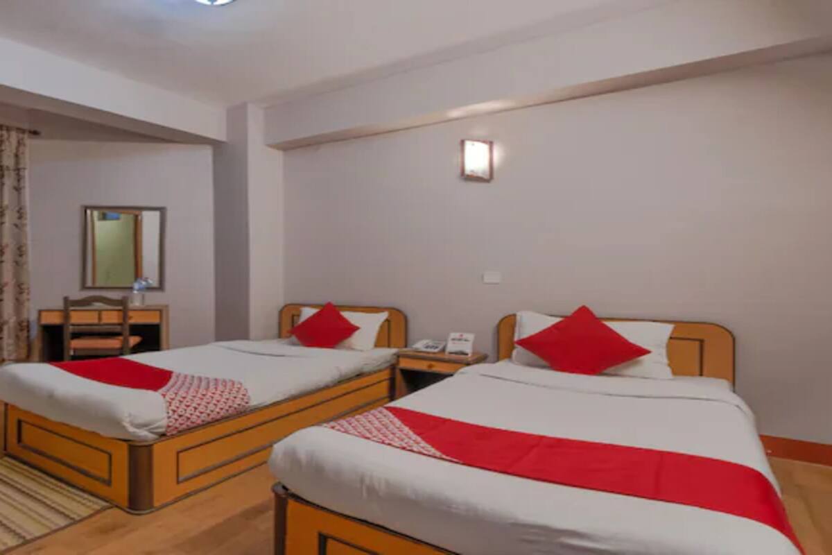 A hotel room with two single beds, each with a red pillow and white bedding. There is a wooden nightstand with a lamp and a telephone between the beds. A mirror hangs on the wall, and a window with curtains is visible.