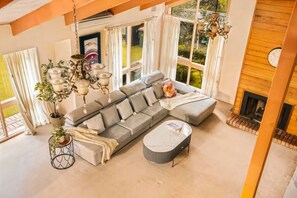 Een moderne woonkamer met een hoekbank, een ronde salontafel, een open haard en een groot raam met gordijnen.