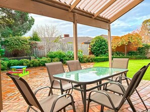 Une terrasse extérieure avec une table et des chaises en verre, entourée d’un sol en briques et d’une clôture en bois.