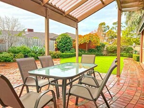 Une terrasse couverte avec une table et des chaises en verre, entourée d’une terrasse en briques et de verdure.