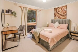 Une chambre à coucher comprenant un lit, des tables de chevet, un bureau, une chaise, une fenêtre avec des rideaux, un tableau accroché au mur et une plante.