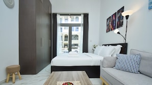 A modern bedroom with a bed, a sofa, a coffee table, a wooden stool, and a view of the outside through a large window. - JVC studio Joya Verde with terrace - 360 vacation (Dubai)