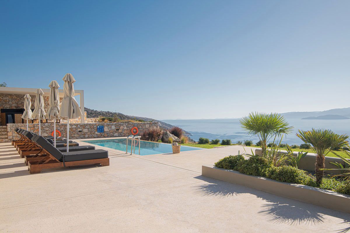 A poolside area with lounge chairs and umbrellas, a stone wall, and a view of the sea.