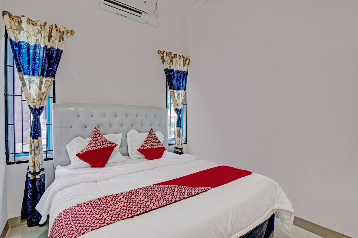 A neatly made bed with white bedding and a red patterned throw, a tufted headboard, and two windows with patterned curtains.