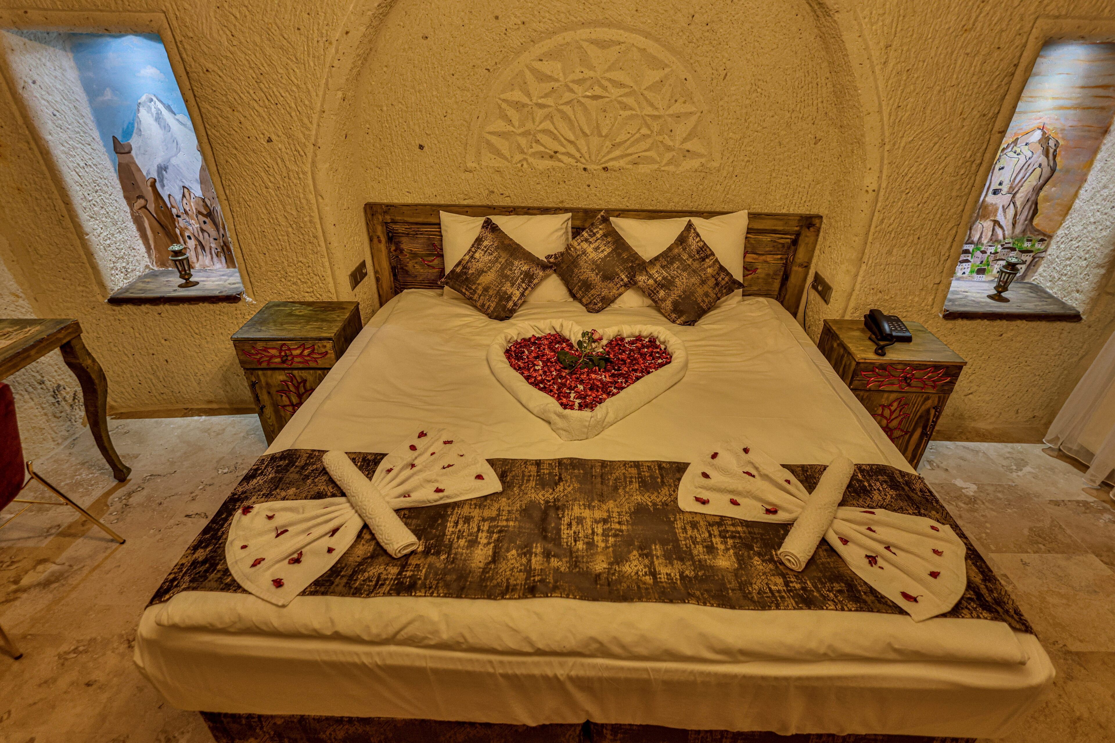 A bed with a heart-shaped arrangement of red flowers, wooden headboard, and two nightstands with a phone and a lamp.