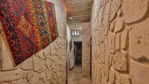 Un corridoio in pietra stretto, con un arazzo decorativo sulla parete sinistra, un soffitto in legno e una scala in fondo.