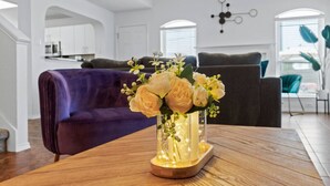 Ein modernes Wohnzimmer mit einem violetten Sofa, einem Couchtisch aus Holz mit einer Blumenvase und einer Küche im Hintergrund.